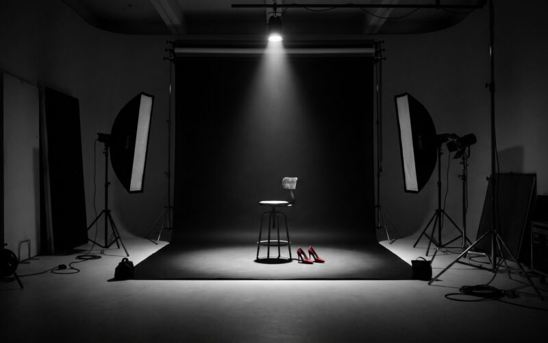 An empty photo studio with a single spotlight on a black backdrop, an empty stool, and a pair of red heels left in the pool of light