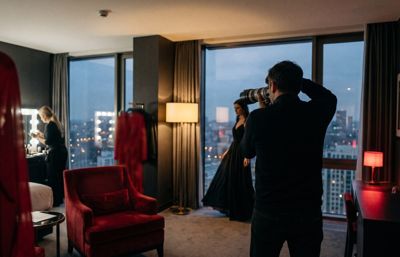 A photographer shooting wide in a dusk-lit hotel suite as a model in a black evening gown poses by floor-to-ceiling windows, with a red velvet chair and a lit makeup station in the room