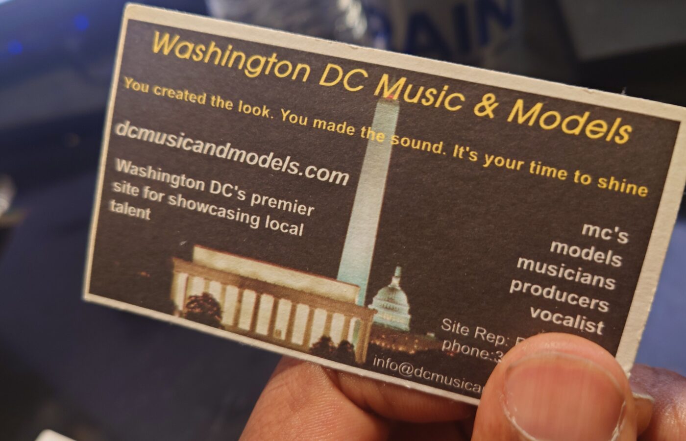 A hand holding the original Washington DC Music & Models business card, printed with the dcmusicandmodels.com address over a night skyline of the Washington Monument and Capitol.