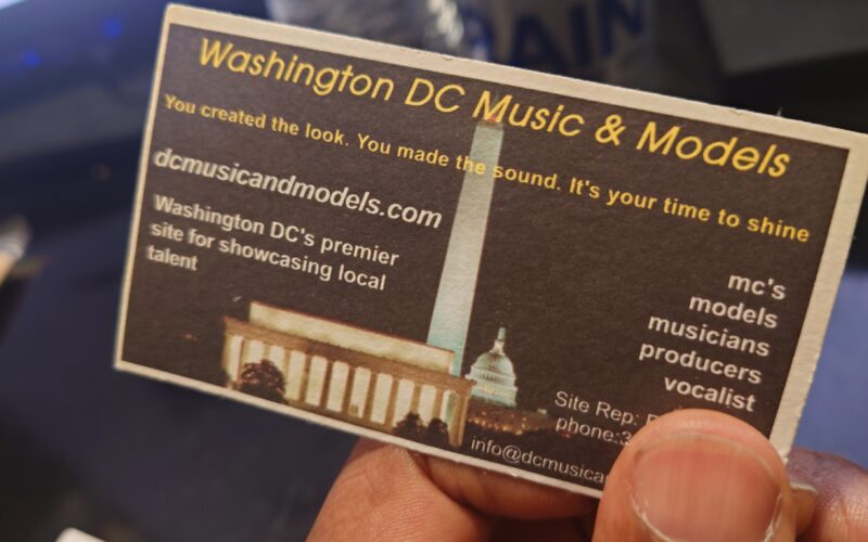 A hand holding the original Washington DC Music & Models business card, printed with the dcmusicandmodels.com address over a night skyline of the Washington Monument and Capitol.