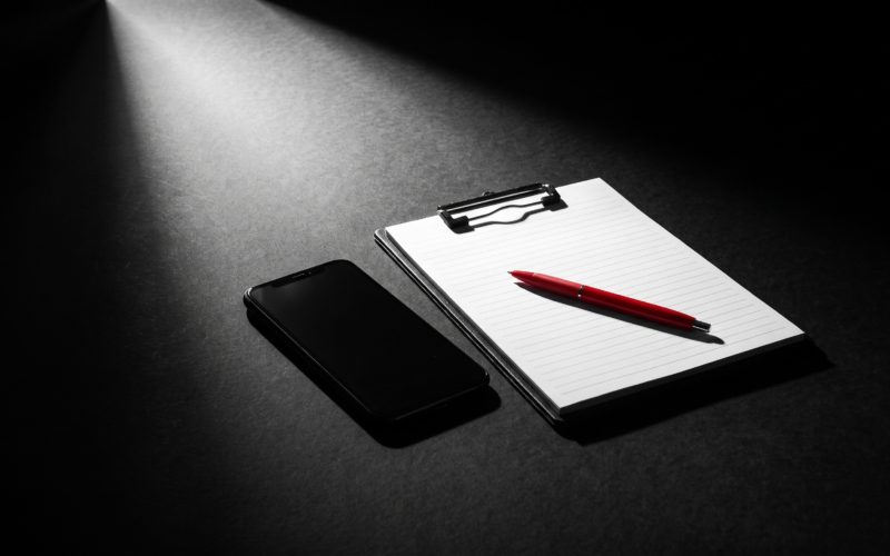 A phone face down on a black studio floor beside a blank shot list on a clipboard with a red pen, under a spotlight