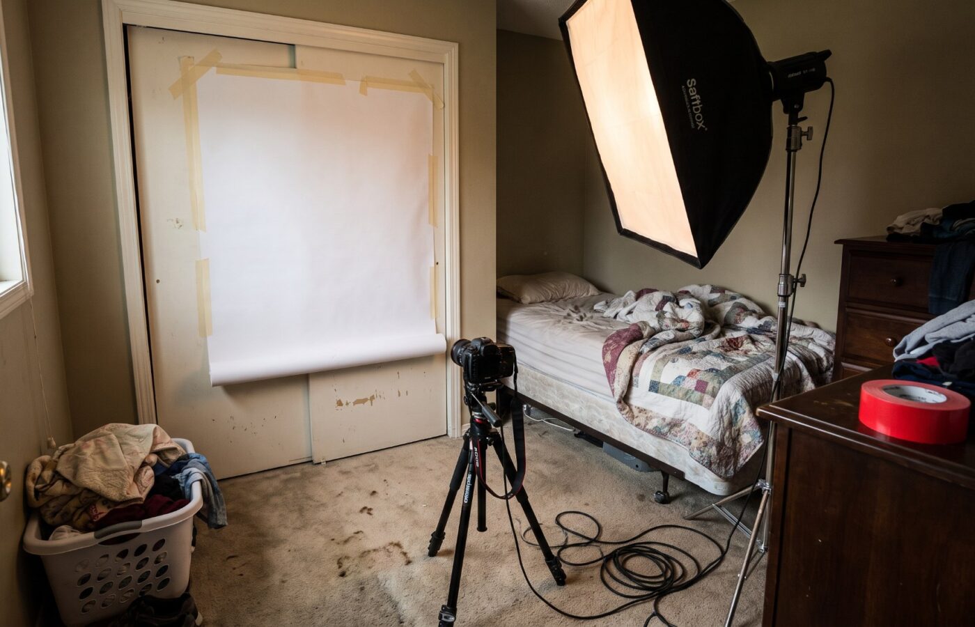 A cramped spare bedroom used as a makeshift photo studio, with seamless paper taped to a closet door and a single softbox wedged beside the bed