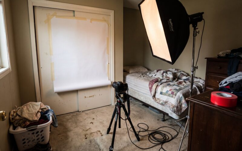A cramped spare bedroom used as a makeshift photo studio, with seamless paper taped to a closet door and a single softbox wedged beside the bed