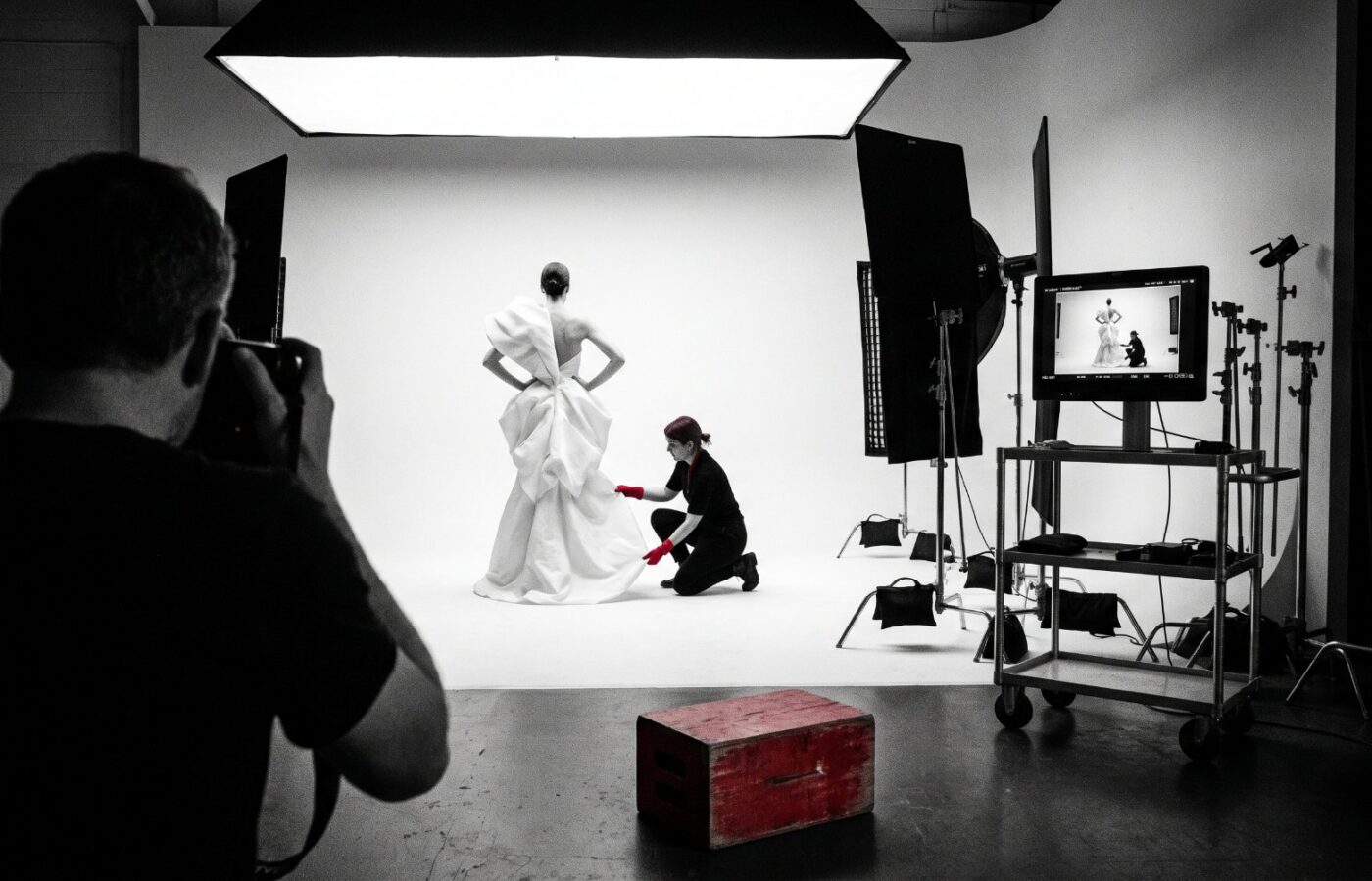 A magazine photoshoot in progress in a dark studio: a model on a white cyclorama under a large overhead softbox, a stylist adjusting the garment, and a tethered monitor on a cart