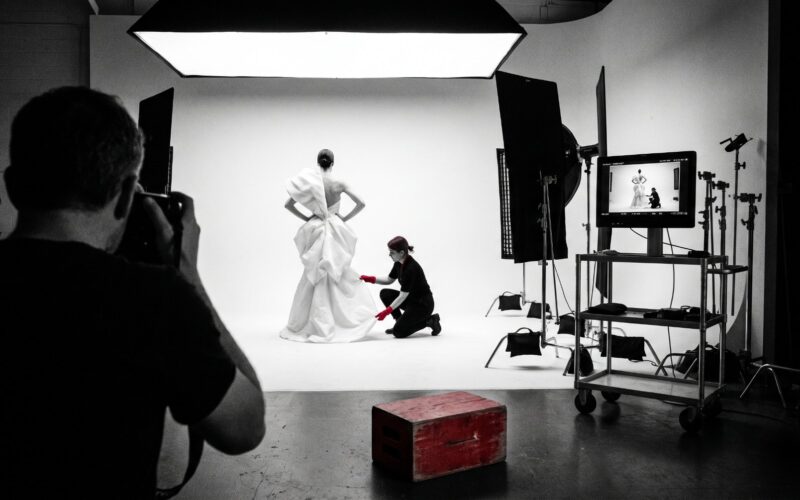 A magazine photoshoot in progress in a dark studio: a model on a white cyclorama under a large overhead softbox, a stylist adjusting the garment, and a tethered monitor on a cart