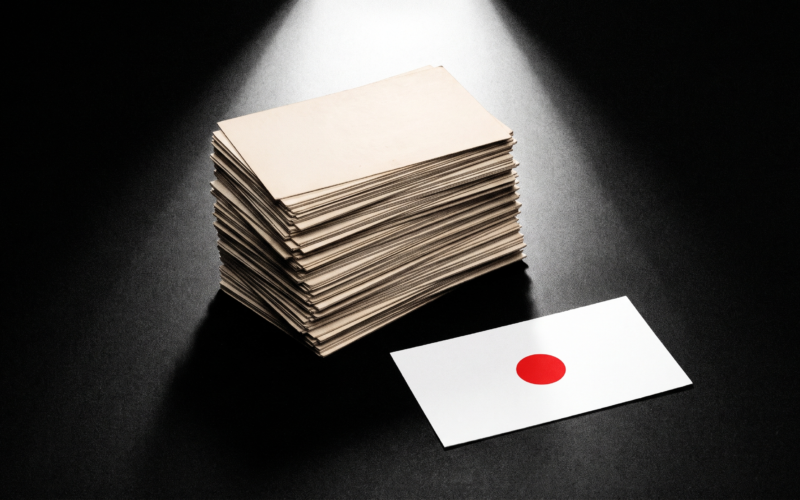 A tall stack of yellowed index cards under a spotlight beside one crisp new card marked with a red dot