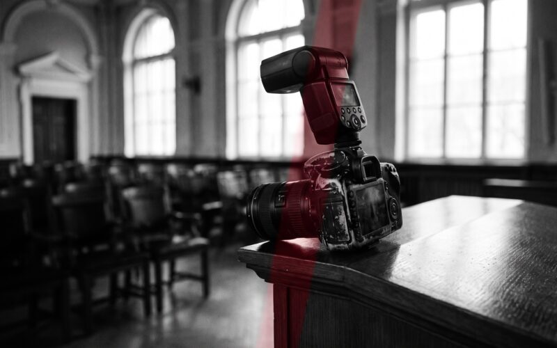 A worn DSLR with an on-camera flash resting on the edge of a podium in an empty ceremony hall, black and white with a red accent light